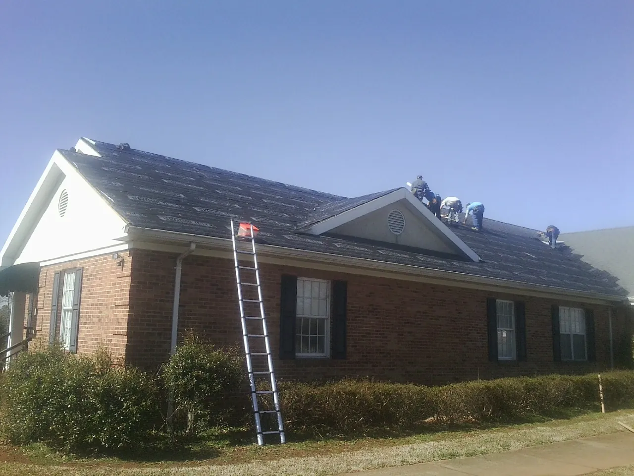 Crew tearing shingles off a brick ranch roof with a ladder set — Emergency Roof Repair work by our crew in Ball Ground