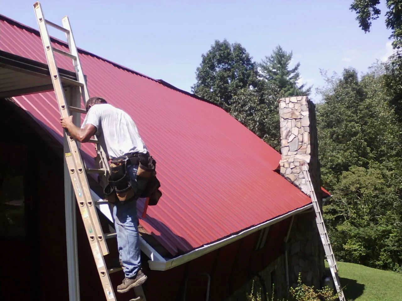 Roofer on a ladder beside a red metal roof and stone chimney — Soffit & Fascia Repair by Roofing Brothers in Ball Ground